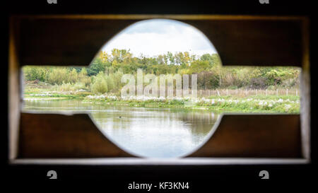 Aussicht von der Vogelbeobachtung Holz- über einem Teich durch eine Öffnung mit einer runden Form geschnitten Hide-out zu Teleobjektive passen. verschwommene Vordergrund (Fenster), shar Stockfoto