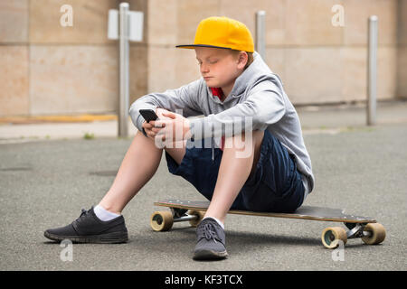 Junge tragen Cap saß auf seinem Skateboard mit Smartphone Stockfoto