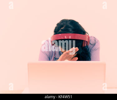 Junge Frau an einem Laptop arbeiten, während das Tragen von Kopfhörern, die ihr Gesicht mit der Hand Stockfoto