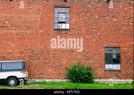 Ein abstraktes Bild von einem verlassenen Auto und alten roten Backstein, verlassenen Lagerhalle Wand mit gebrochenen Quadratischen Fenstern. Stockfoto