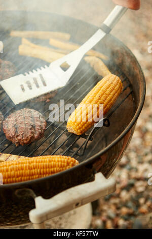 Person kochen Fleisch und Maiskolben zum Grillen Sie Grillen über den heißen Kohlen mit einem Metallspatel in der Nähe zu sehen. Stockfoto