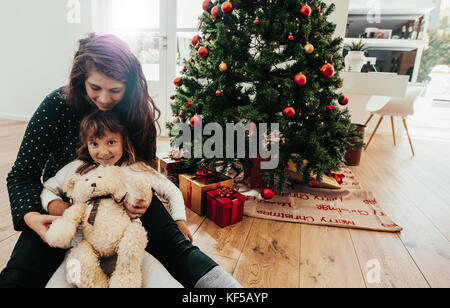 Kleines Mädchen mit ihrer Mutter mit einem Teddybär in Weihnachten. Schön Weihnachtsbaum mit geschenkboxen eingerichtet. Stockfoto