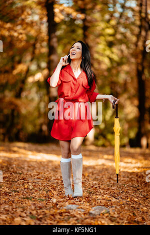 Glückliche junge Frau im roten Kleid sprechen auf dem Smartphone Wandern im Herbst sonnigen Park, Holding gefaltet Yellow Umbrella. Stockfoto