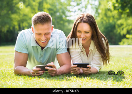 Junges Paar Liegen auf Gras auf der Suche auf ihre Handys in Park Stockfoto