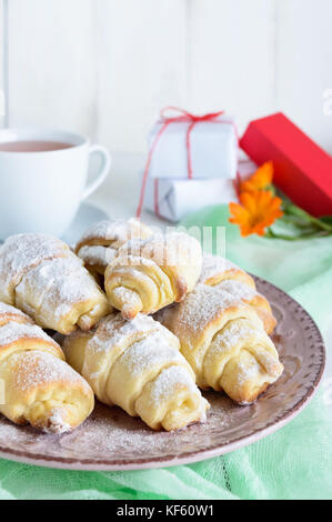 Hausgemachte Croissants mit Marmelade, dekoriert mit Puderzucker auf einer Keramikplatte und Tasse Tee am Morgen auf einem weißen Holz- Hintergrund. Stockfoto