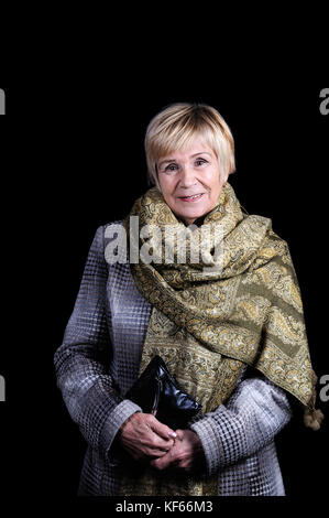 Portrait von einer älteren Frau in einem Drapierten Mantel mit einem geworfenen Schal auf ihre Schultern und Clutch. schwarzen Hintergrund. Stockfoto