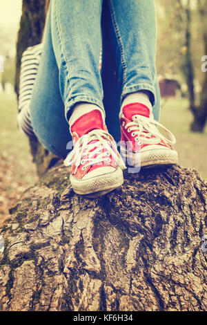 Die Beine der Frau das Tragen der roten canvas Turnschuhe und Jeans, sitzt auf einem Baum, im Retrostil Foto Stockfoto