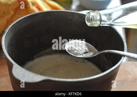 Prozess der Abschrecken der Natron mit dem Essig in einem Löffel über eine gusseiserne Schüssel mit dem Teig. close-up Bild. Prozess des Kochens. Stockfoto