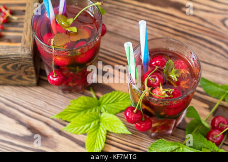 Gläser erfrischenden Drink gewürzt mit frischem Obst und mit Kirschen mit Tautropfen bedeckt gehalten. Holz- Hintergrund Stockfoto