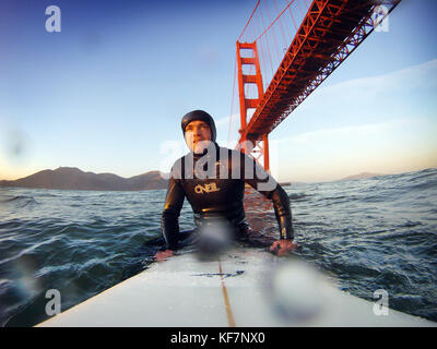 Usa, Kalifornien, San Francisco, Fort Point, Surfer sitzen auf seinem Surfboard unter der Golden Gate Bridge bei Sonnenuntergang Stockfoto