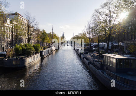 Am späten Nachmittag scheint die Sonne über einem Kanal, der von Hausbooten und klassischer Architektur gesäumt ist, in einer belebten Stadt. Stockfoto