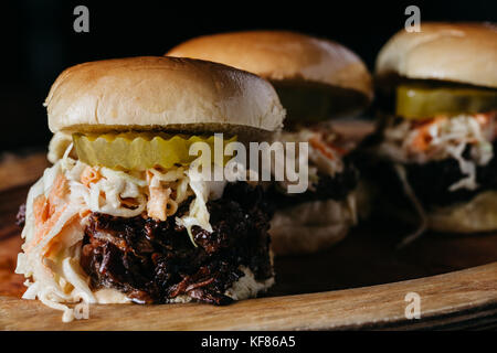 Schieber mit gezogenen Schweinefleisch, Essiggurken und Cole Slaw auf einer Holzplatte mit dunklen Hintergrund Stockfoto