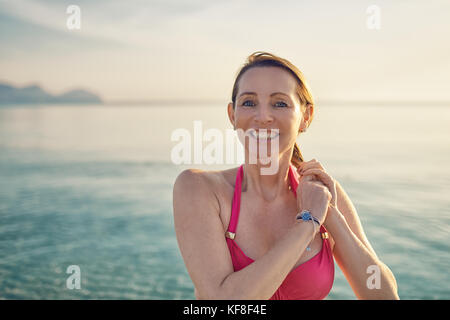 Glücklich lächelnde Frau mittleren Alters das Festhalten an ihr langes Haar stand mit dem Rücken zum Meer und Glühen der Sonne am Meer, als sie einen Tag genießt Stockfoto