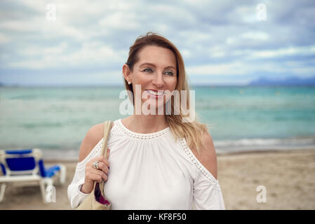 Attraktive trendy blonde Frau in einem stilvollen oben stehend auf einem tropischen Strand mit Blick auf die Kamera mit einer freundlichen und entspannten Lächeln mit ihr zurück t Stockfoto