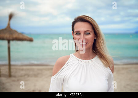 Attraktive trendy blonde Frau in einem stilvollen oben stehend auf einem tropischen Strand mit Blick auf die Kamera mit einer freundlichen und entspannten Lächeln mit ihr zurück t Stockfoto