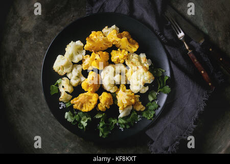 Weißer und gelber Blumenkohl Gratin auf schwarze Keramikplatte mit Korianderblättern, Textil, Servietten und Besteck über Dark Metal Textur Hintergrund serviert. Stockfoto