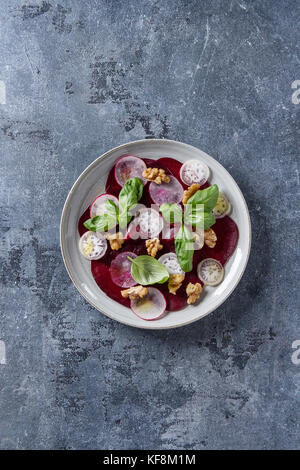 Vegan rote Beete carpaccio Salat mit Radieschen, Basilikum, Olivenöl und Nüssen in Keramik Platte über blaue Textur Hintergrund. top Aussicht, Raum. Gesunde eatin Stockfoto