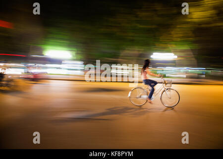 VIETNAM, Hanoi, eine junge Frau fährt nachts mit dem Fahrrad eine belebte Straße entlang, den Hoan Kiem See in der Ferne Stockfoto
