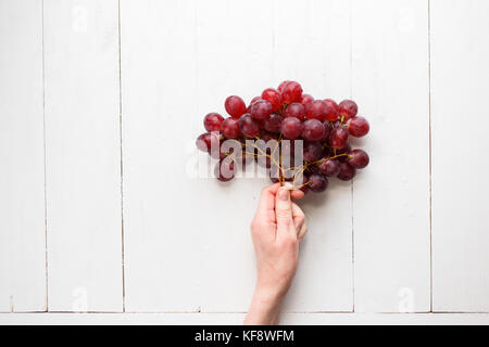 Die Hand des Mädchens hält ein Bündel von roten Trauben auf einem hölzernen Hintergrund. Blick von oben. Trauben sind wie Luftballons. Stockfoto