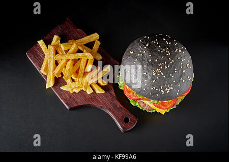 Schwarz Burger mit Pommes frites auf Holz Schneidebrett auf schwarzem Hintergrund isoliert. oben flach. Stockfoto