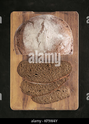 In Scheiben geschnittene hausgemachte Sauerteig Roggenbrot auf Schneidebrett über schwarzen strukturierten Hintergrund mit Roggenmehl. Ansicht von oben oder flach-legen. Low Key Stockfoto