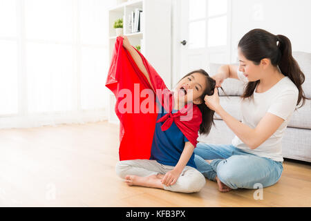 Junge lächelnde Mutter helfen, kleine Mädchen gebunden Haar im Wohnzimmer, wenn Tochter als Superheld zu Hause mit rotem Umhang spielen möchten. Stockfoto