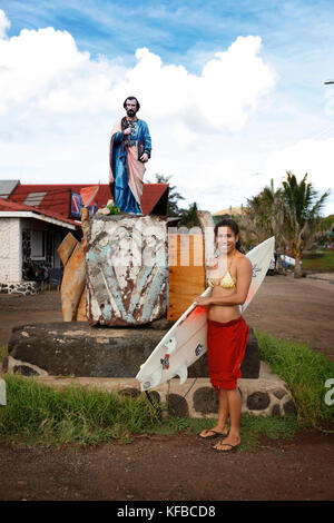 Easter Island, Chile, Isla de Pascua, Rapa Nui, eine Frau steht mit ihrem Surfbrett in der Nähe einer Statue im Herzen der Innenstadt von Hanga Roa Stockfoto