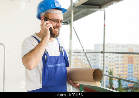 Lächelnd Bauarbeiter in Outfit und in Helm stehen in großer Höhe auf einer Baustelle mit Plänen unter den Arm und Reden auf t Stockfoto