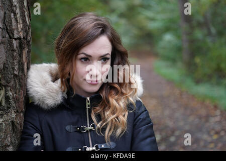 Nachdenkliche junge Frau lehnte sich gegen Baum im Wald Stockfoto