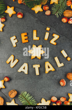 FELIZ NATAL COOKIES. Wörter Frohe Weihnachten en Portugiesisch mit gebackenen Plätzchen, Weihnachtsschmuck und Muttern auf schwarzem Schiefer Hintergrund. Christmas Card f Stockfoto