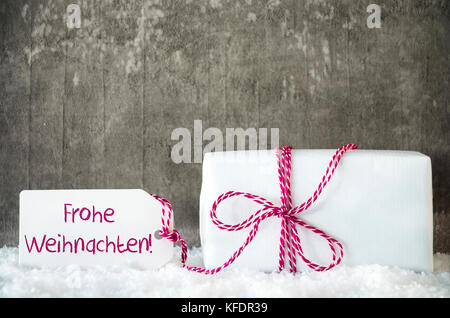 Eine weiße Geschenk mit Label mit deutschem Text frohe Weihnachten bedeutet, frohe Weihnachten. Grau grungy Zement Hintergrund mit Schnee. Stockfoto