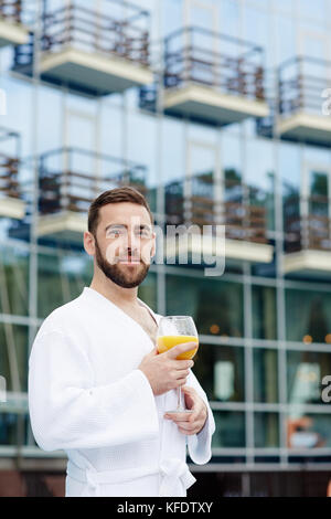 Ruhiger junger Mann mit einem Glas Getränk, der einen weißen Bademantel trägt, während er sich entspannt Stockfoto
