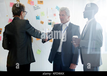 Gutaussehender Senior Entrepreneur im klassischen Anzug, der seiner Geschäftspartnerin nach erfolgreichem Abschluss der Verhandlungen die Hand schüttelt, African A Stockfoto