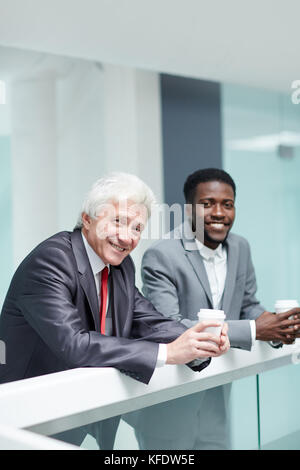 Der gutaussehende junge Manager und sein Senior Chef posieren für Fotografie mit einem lächelnden Lächeln und genießen duftenden Kaffee in der modernen Bürolobby Stockfoto