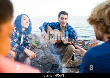 Glückliche Kerl mit Gitarre und seinen Freunden zu verbringen um am Abend am Lagerfeuer von Seaside Stockfoto