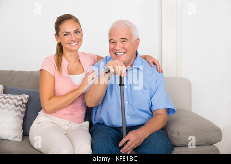 Lächelnde junge Frau mit ihrem behinderten Vater sitzt auf einem Sofa zu Hause Stockfoto