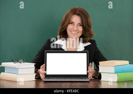 Glückliche junge Lehrerin mit Laptop im Klassenzimmer Schreibtisch sitzen Stockfoto