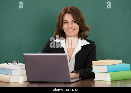 Junge Lehrerin mit Laptop am Schreibtisch im Klassenzimmer sitzen Stockfoto