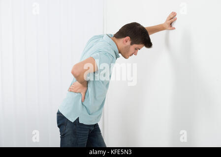 Junge Mann in Schmerz in seinem Rücken, während gegen die Wand zu Hause stehend Stockfoto