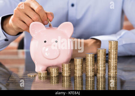 Geschäftsmann Münzen einfügen in Sparschwein mit Stapel von Münzen über den Schreibtisch Stockfoto