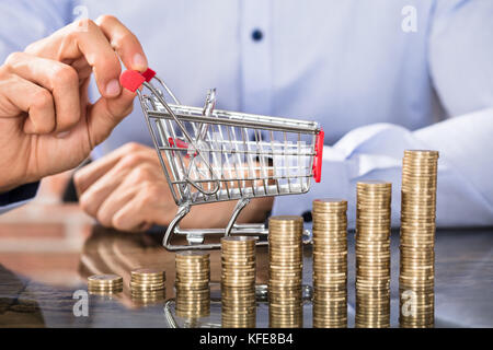 Stapel von zunehmenden Münzen vor dem Geschäftsmann holding Einkaufswagen auf Schreibtisch Stockfoto