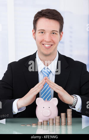 Portrait von selbstbewussten jungen Geschäftsmann schützenden Sparschwein und Münze Stapel im Büro Schreibtisch Stockfoto