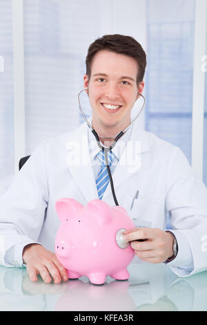 Portrait von jungen männlichen Arzt untersuchen Sparschwein mit Stethoskop im Krankenhaus Stockfoto