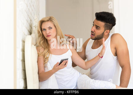 Junges Paar sitzen im Bett, Frau mit Zelle smart phone Hispanic Mann frustriert, Problem Stockfoto