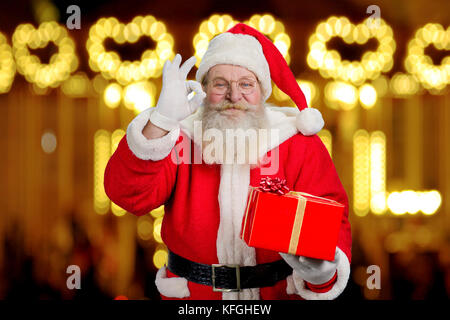 Smiling Santa Claus mit okey unterzeichnen. Santa Claus mit echtem Bart holding Weihnachtsgeschenk und okey Geste mit Fingern auf backgroun verschwommen Stockfoto