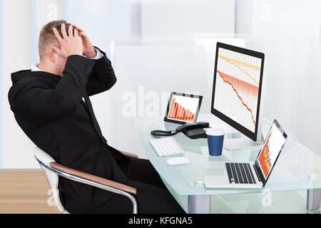 Verzweifelten Geschäftsmann mit finanziellen Verlusten an seinem Schreibtisch consulting drei Graphen auf verschiedenen Monitoren alle fallen in die roten konfrontiert Stockfoto