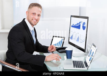 Geschäftsmann mit guter Performance graph an seinem Schreibtisch mit statistischen Berichte über drei Monitore Jubel und mit seinen Fäusten umklammerte verteilt sitzen Stockfoto