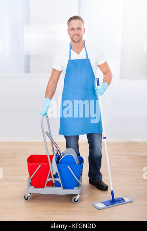 Hausmeister Reinigung Parkett mit einem Mopp und eine Karre mit zwei Eimer für das Desinfektionsmittel und Wasser anhalten zu lächeln in die Kamera, während er geht über h Stockfoto