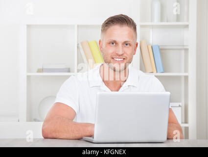 Lächelnder junge Mann in ein kurzes sleeved Hemd sitzt zu Hause auf einem Laptop im home Office arbeiten Stockfoto