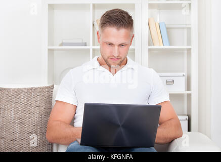 Attraktive junge Mann sitzt in seinem Wohnzimmer auf dem Sofa mit einem Laptopcomputer ausgewogen auf seinem Knie zum Surfen im internet Stockfoto